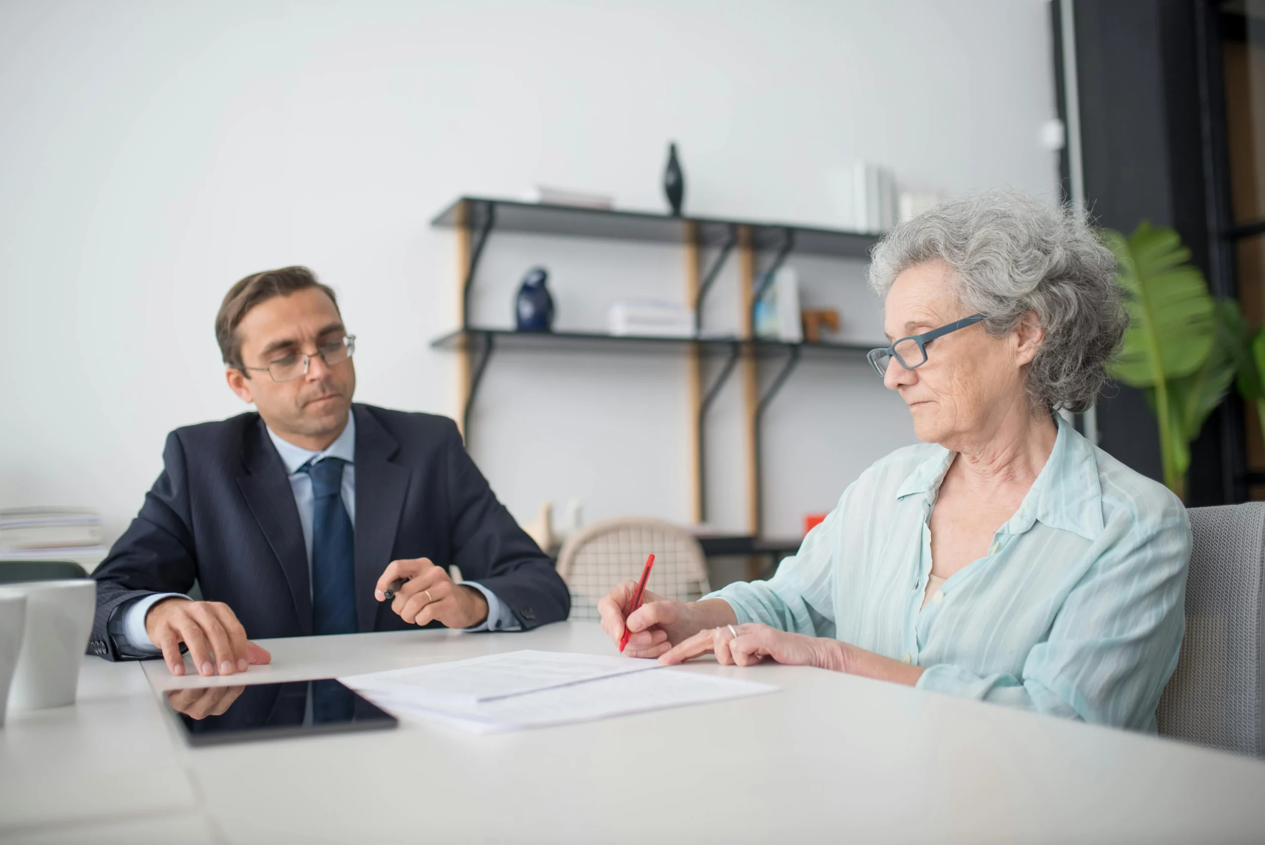 Une femme sénior signant un document avec un stylo rouge lors d'un rendez-vous avec un conseiller funéraire en costume
